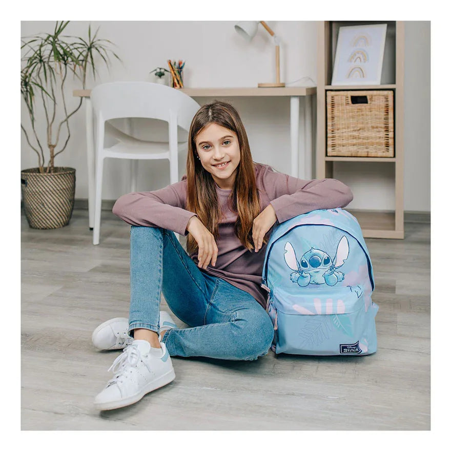 Menina sorrindo sentada ao lado de uma mochila infantil azul do Stitch em ambiente doméstico